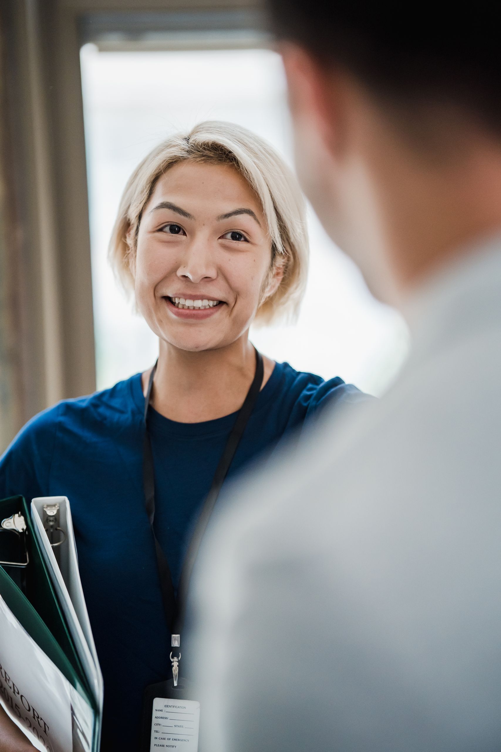 A cheerful woman in a professional setting holds binders, smiling at a coworker.